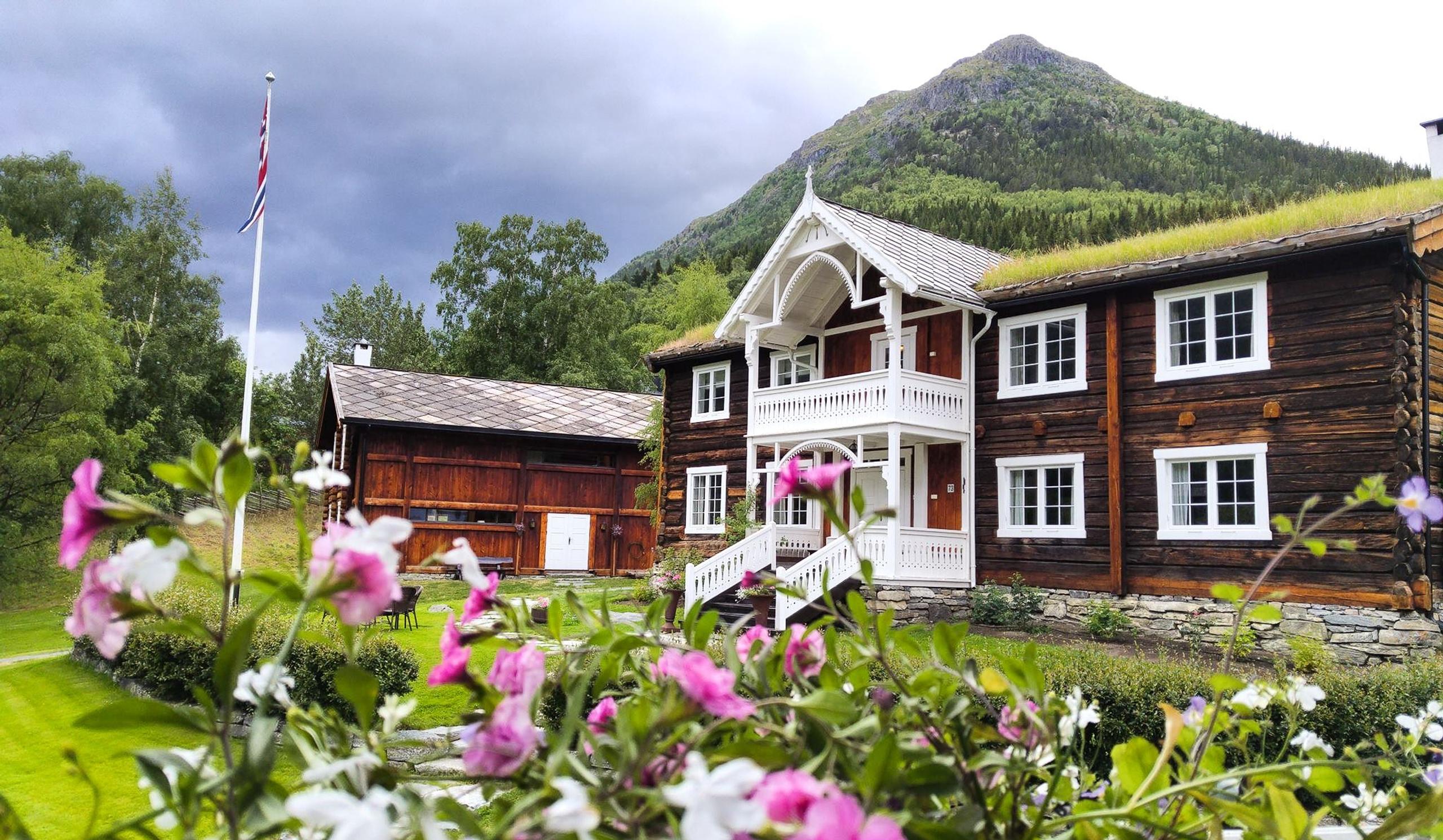 Tømmerhus med kvit veranda og torvtak, blomstrar i forgrunnen, fjell og mørke skyer i bakgrunnen..