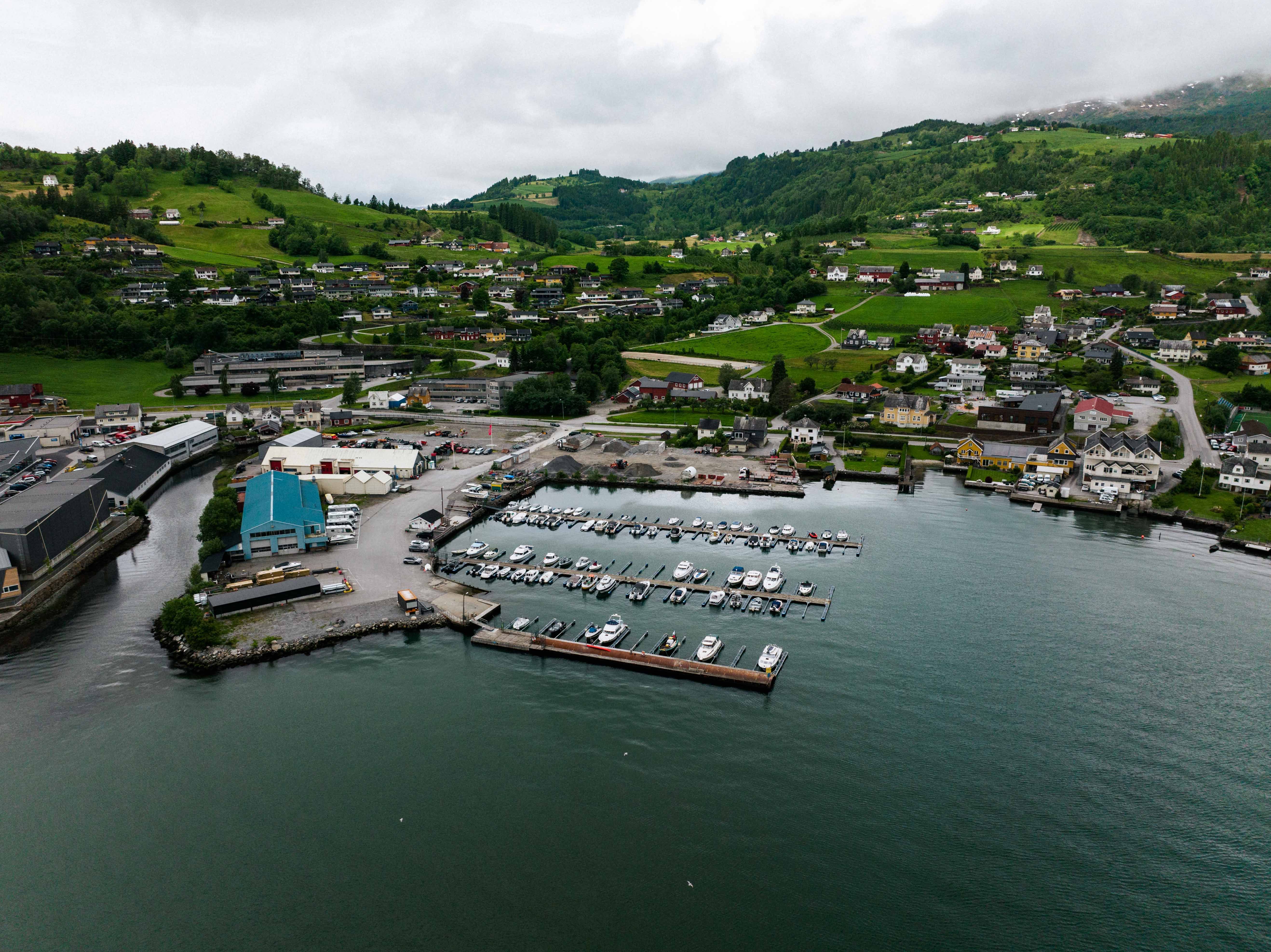 Panoramic aerial view of Øystese Guest Harbour and the village, showing the Hardangerfjord, green hills, and boats in the marina.
