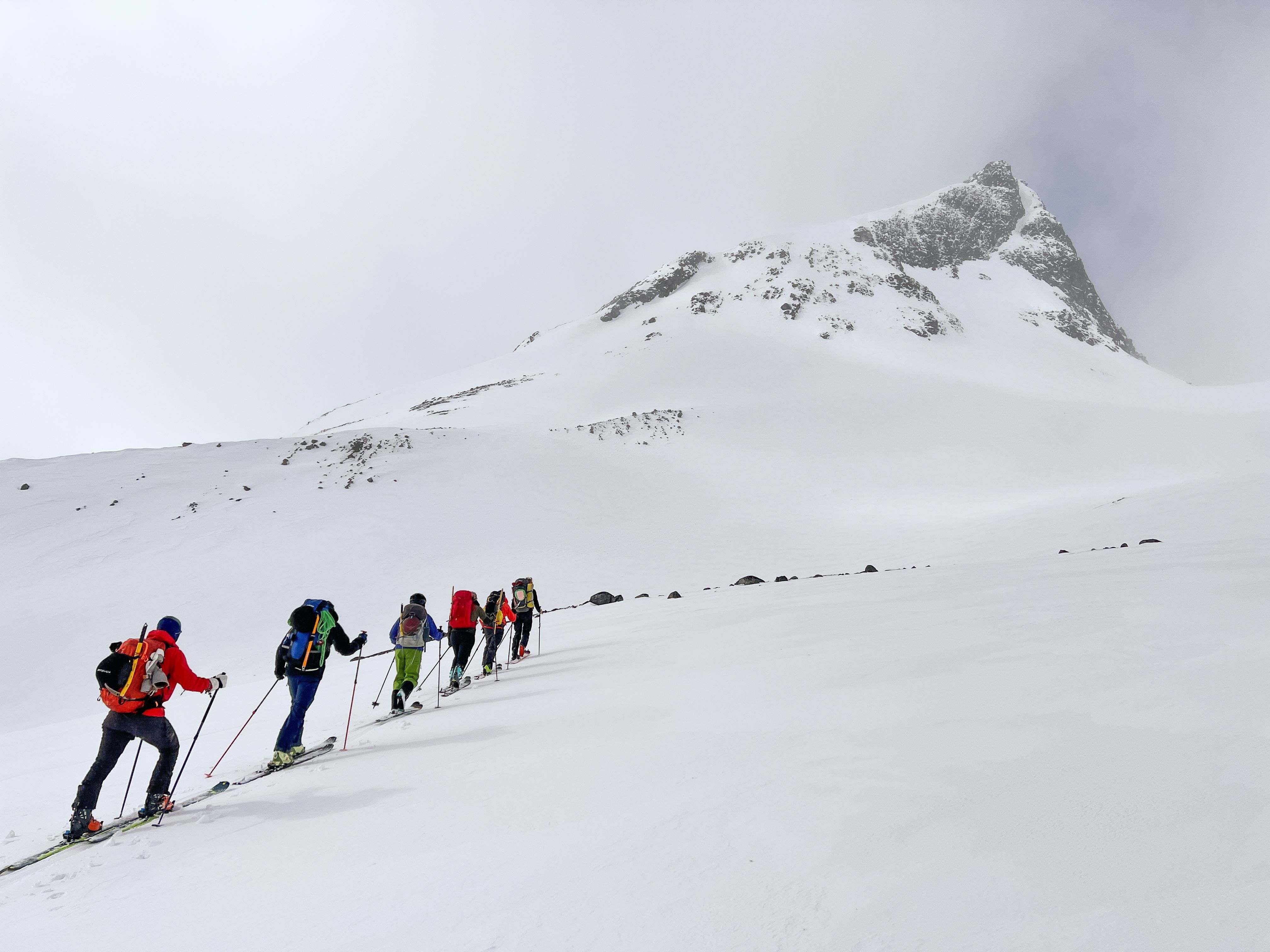 Groep backcountry-skiërs met rugzakken en stokken die een besneeuwde helling beklimmen naar een rotsachtige top onder wolken..
