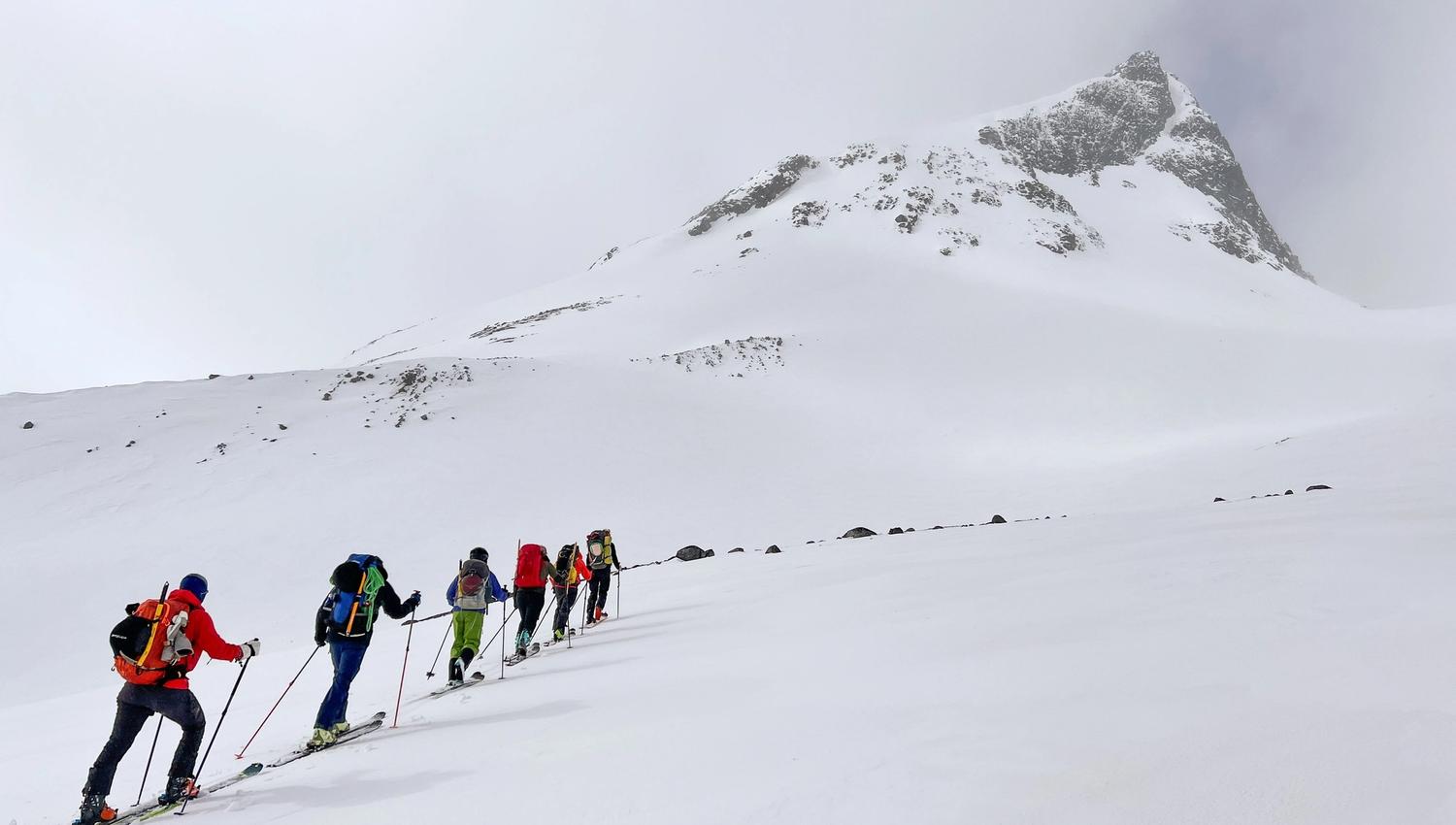 Groep backcountry-skiërs met rugzakken en stokken die een besneeuwde helling beklimmen naar een rotsachtige top onder wolken..