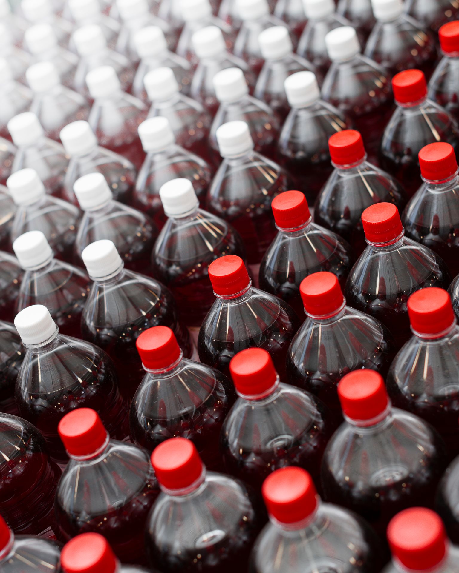 Mange plastflasker med mørk brus, toppet med røde og hvite korker, ordnet i rader.