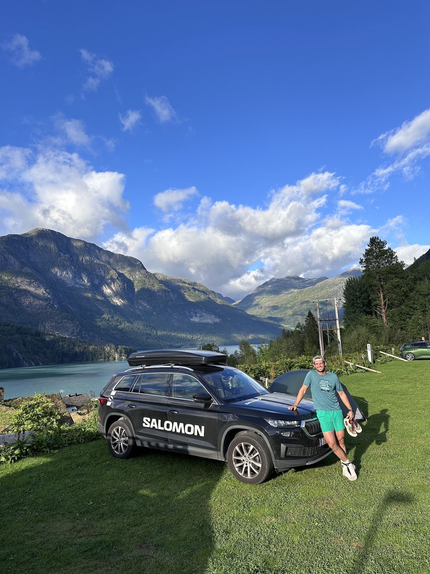 Person leaning on a dark SUV on a grassy hill, with a lake, forested mountains, and blue sky with clouds..