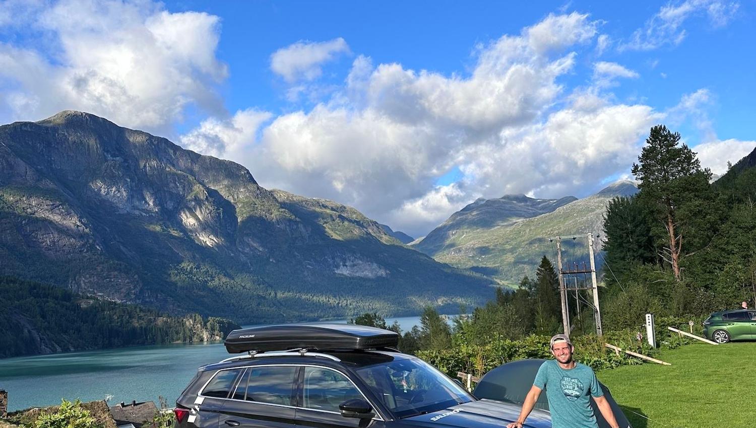 Person leaning on a dark SUV on a grassy hill, with a lake, forested mountains, and blue sky with clouds..