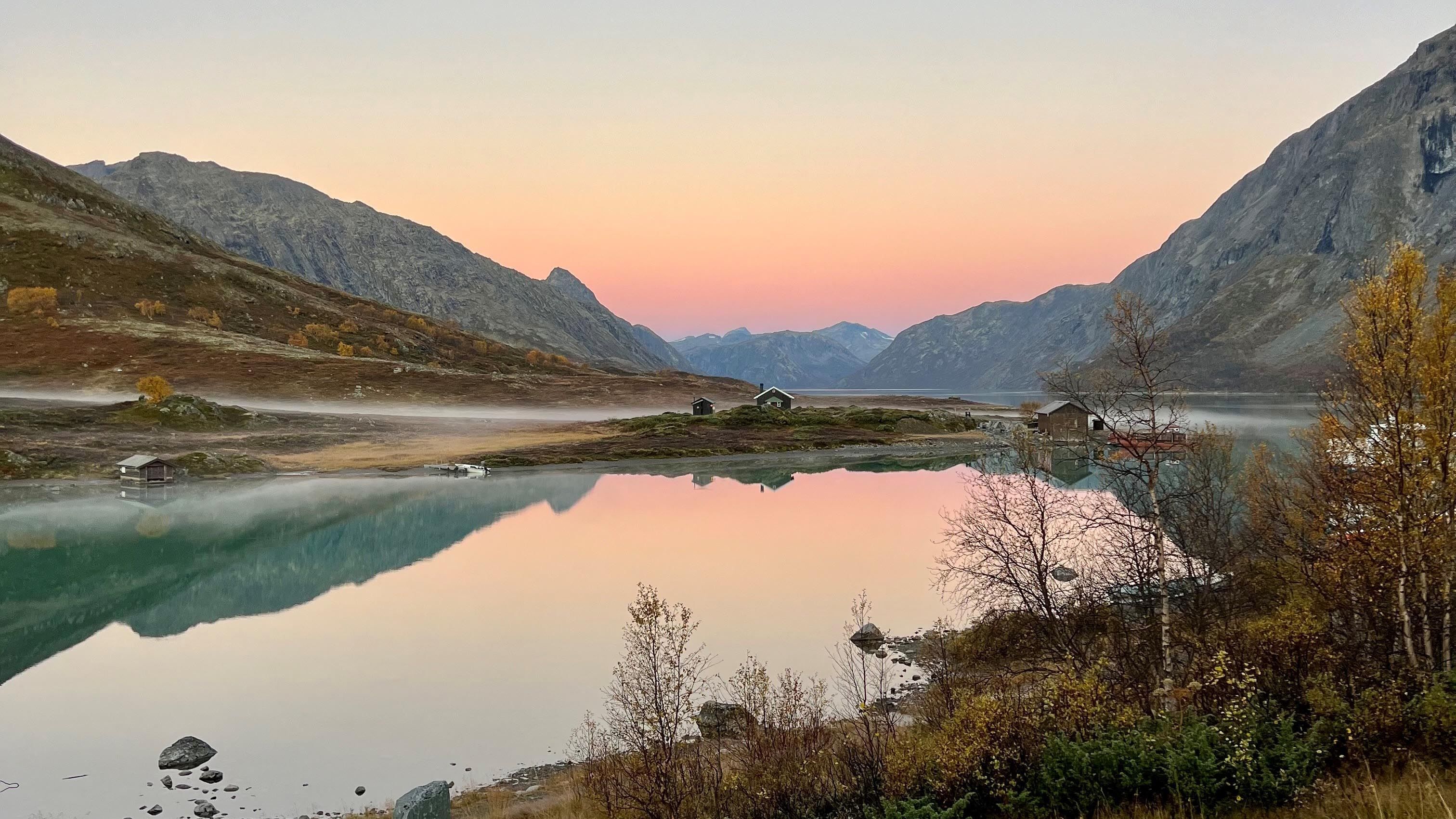 Calm mountain lake at sunrise with pastel sky, misty shore, small cabins, and rocky slopes reflected in water..