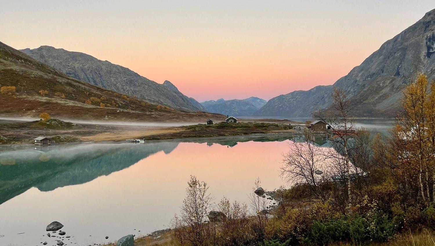 Calm mountain lake at sunrise with pastel sky, misty shore, small cabins, and rocky slopes reflected in water..