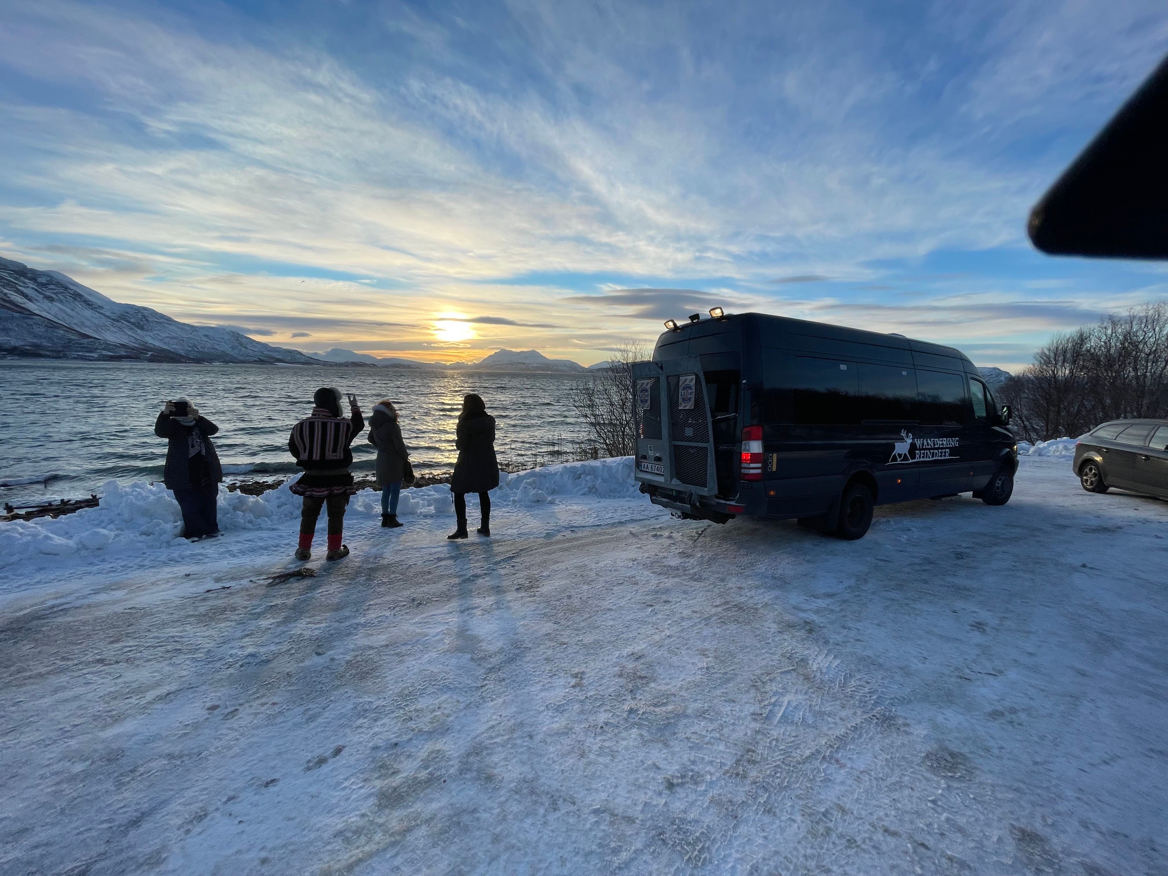 People at a snowy viewpoint with fjord, mountains, and sunset