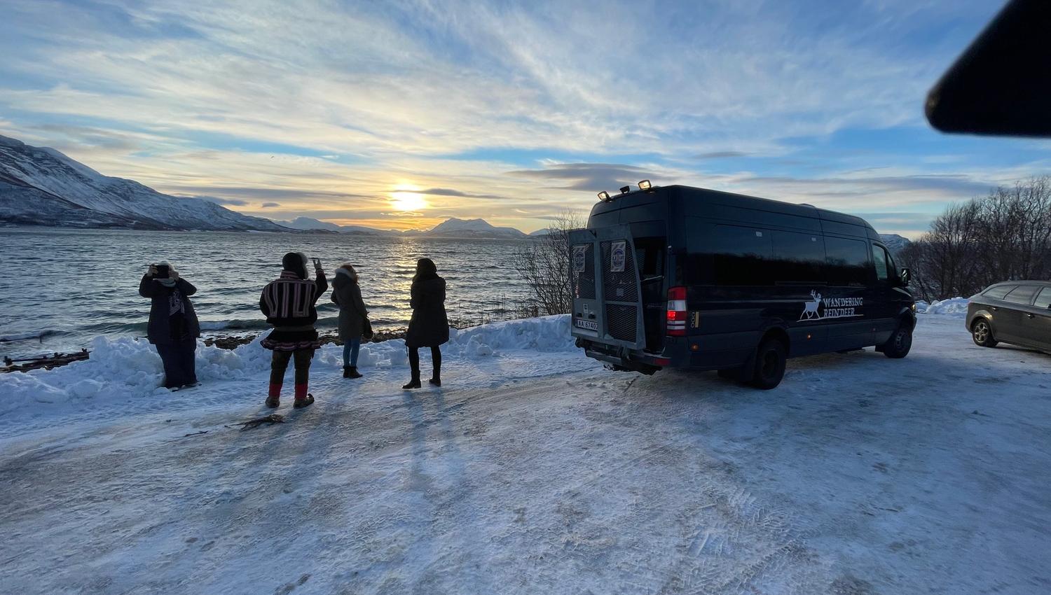 People at a snowy viewpoint with fjord, mountains, and sunset
