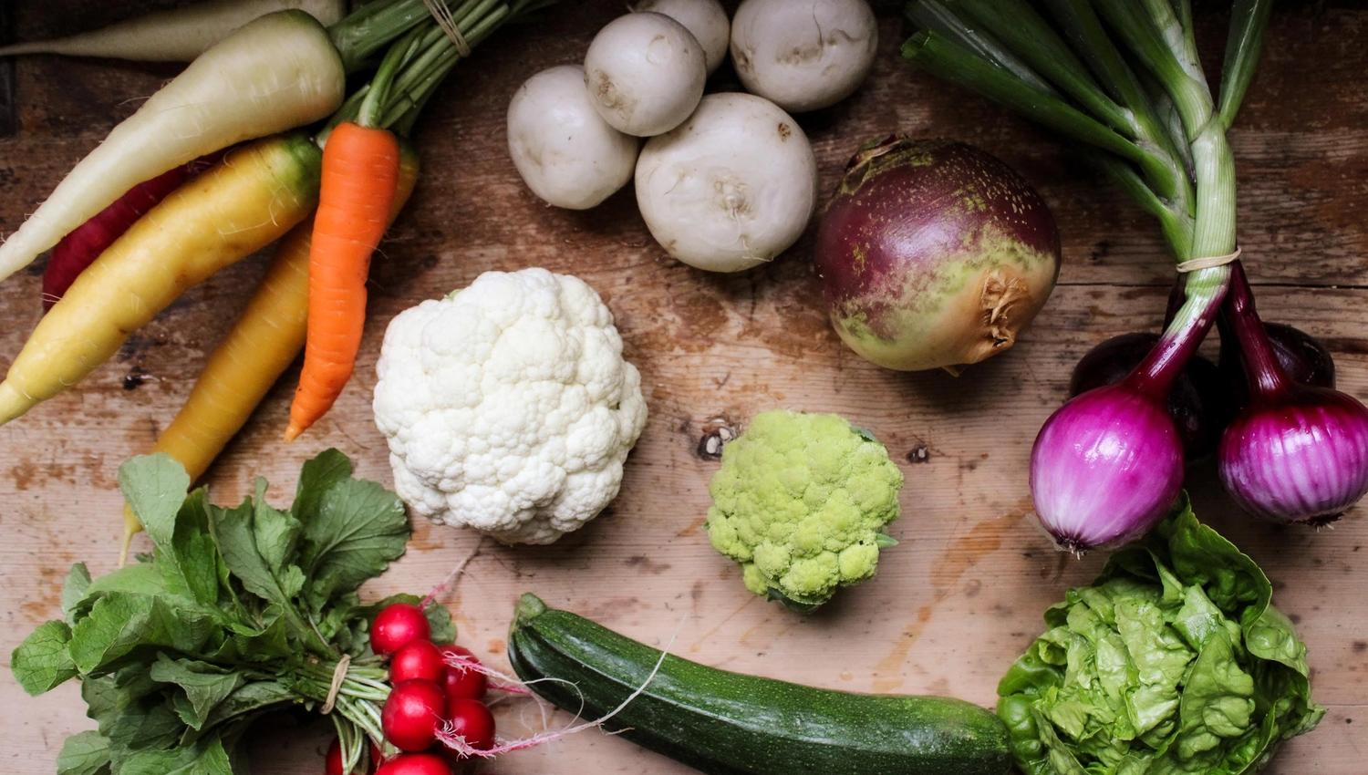 Top-down view of assorted fresh vegetables on a rustic wooden surface, including carrots, onions, radishes, and cauliflower..