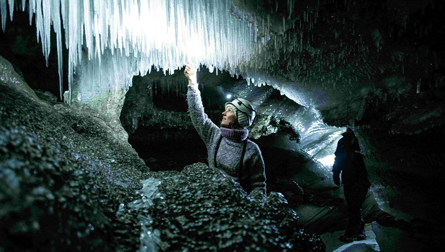 En guide med en lys hodelykt og hjelm på inne i en isgrotte som kjenner på en istapp i taket av grotten
