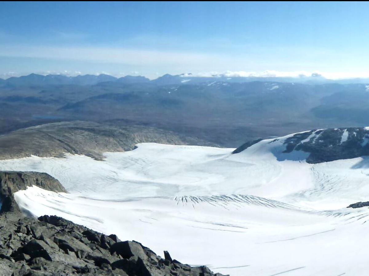 View of a wide glacier in a mountain valley, with rocky foreground, distant mountain ridges and clear blue skies.