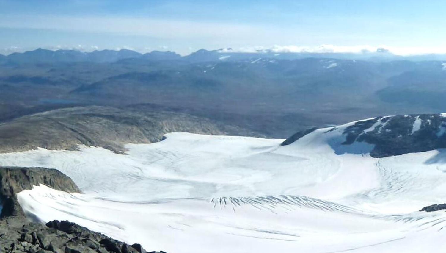 View of a wide glacier in a mountain valley, with rocky foreground, distant mountain ridges and clear blue skies.