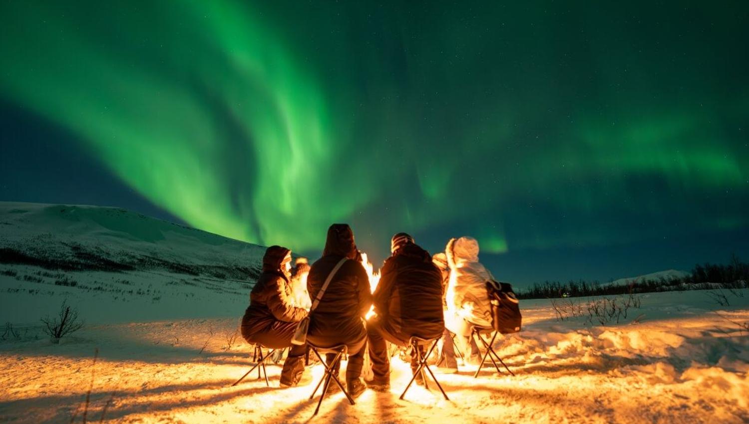 Guests cozy around a fire while the Northern Lights dance overhead