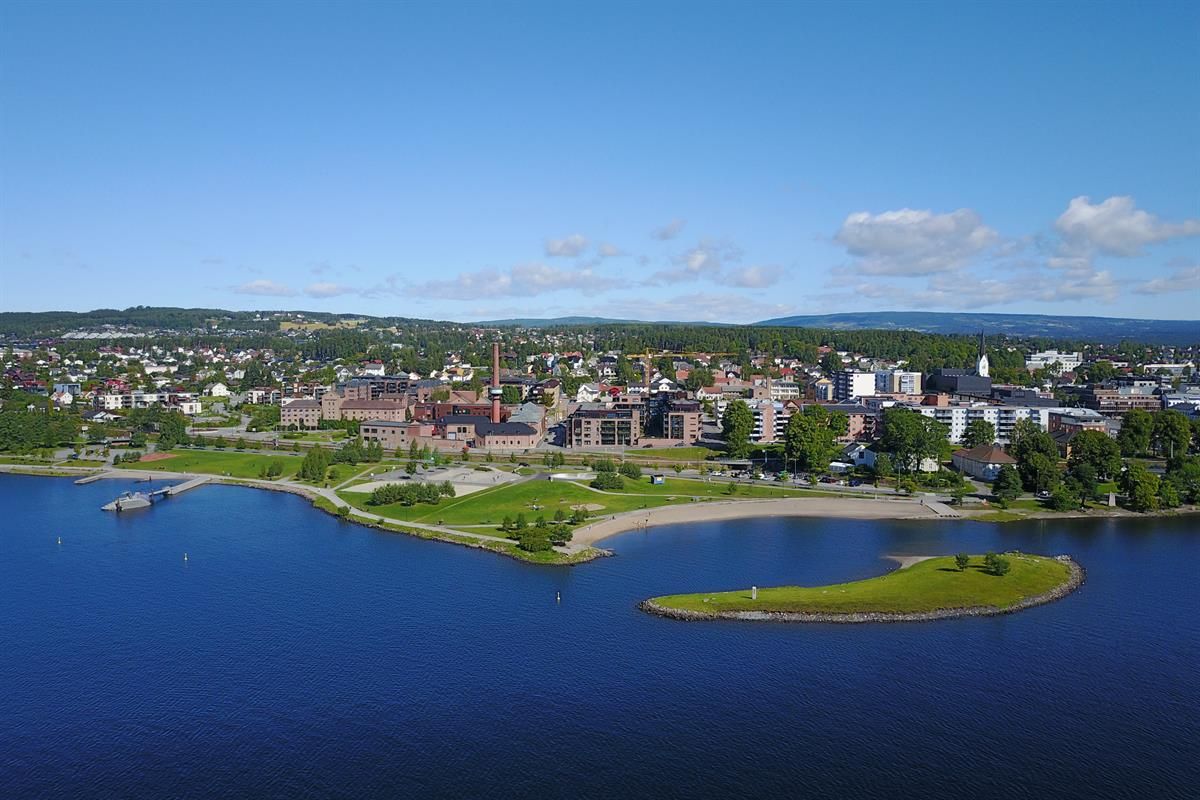 Oversiktsbilde av en by ved et stort vann, med grønt landskap og spredte skyer på himmelen.