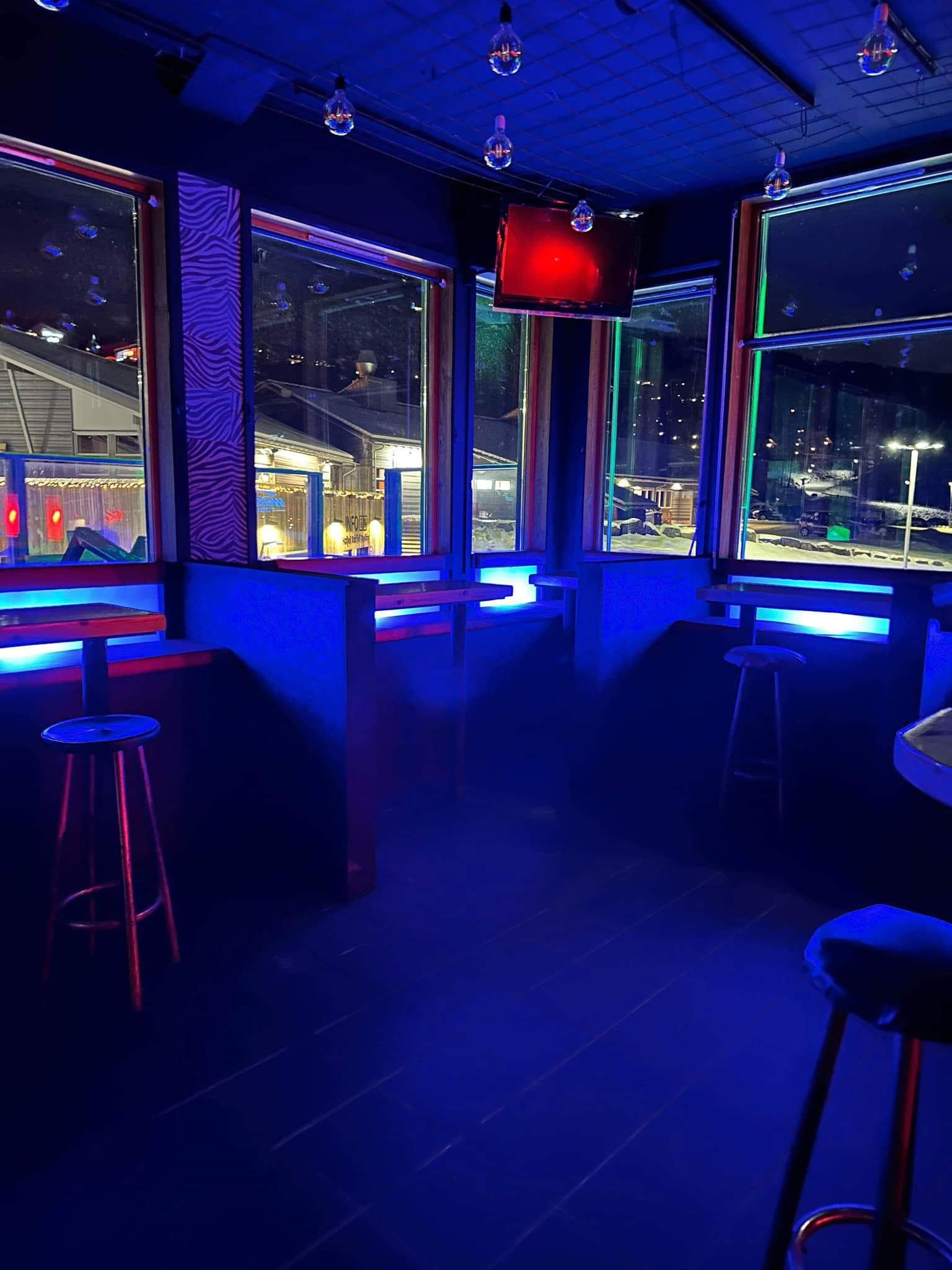 Tomt barhörn med barstolar, bord och stora fönster mot en upplyst vinterkväll, blå neon och tv-skärm.