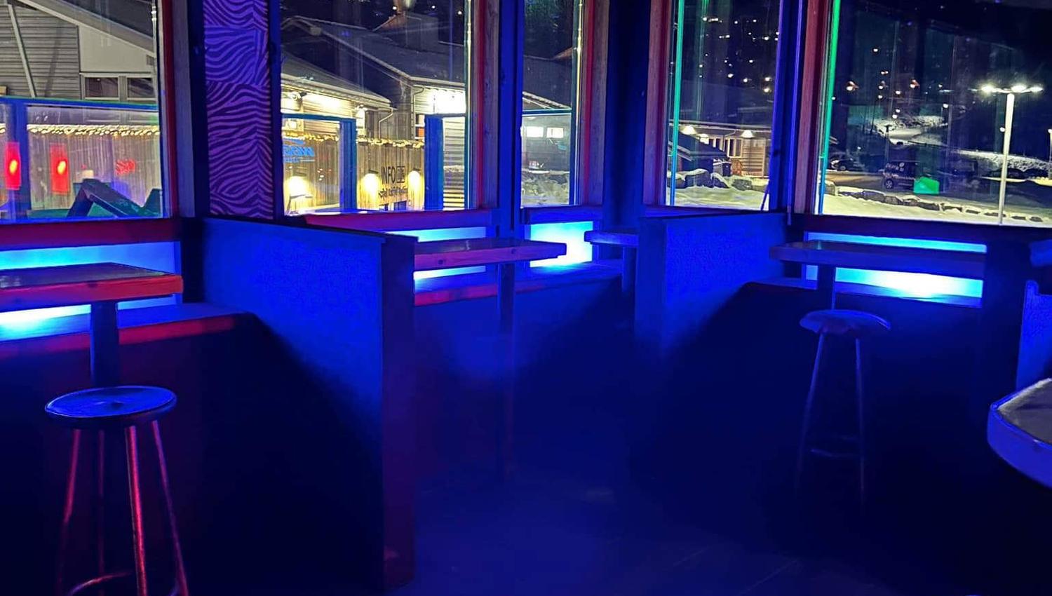 Tomt barhörn med barstolar, bord och stora fönster mot en upplyst vinterkväll, blå neon och tv-skärm.