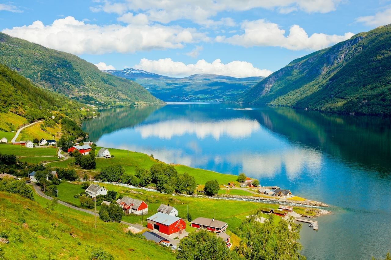 Kandal, Gloppen - Green farmland with red and white houses by a calm fjord, surrounded by mountains under a blue sky with clouds..
