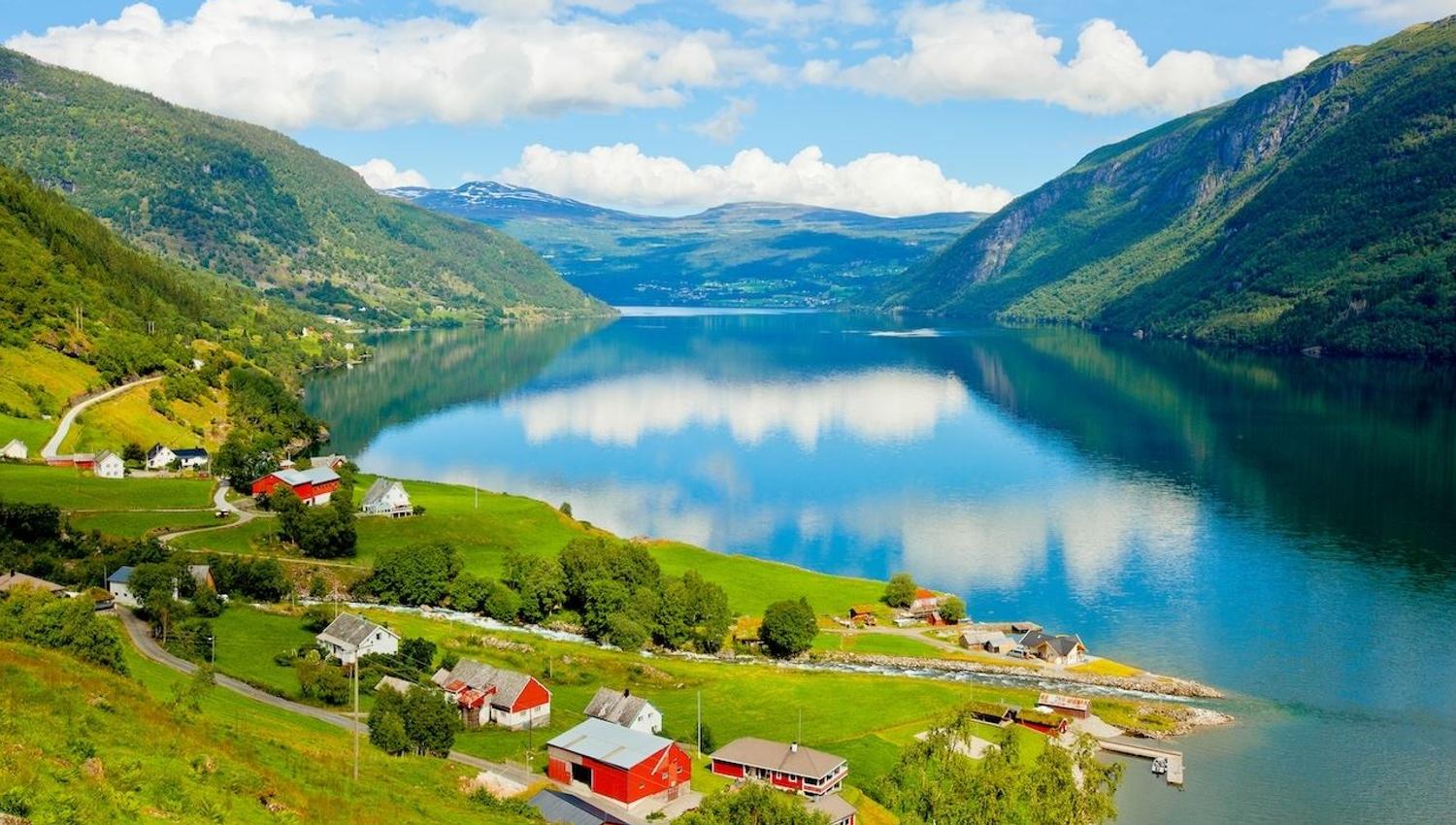 Kandal, Gloppen - Green farmland with red and white houses by a calm fjord, surrounded by mountains under a blue sky with clouds..