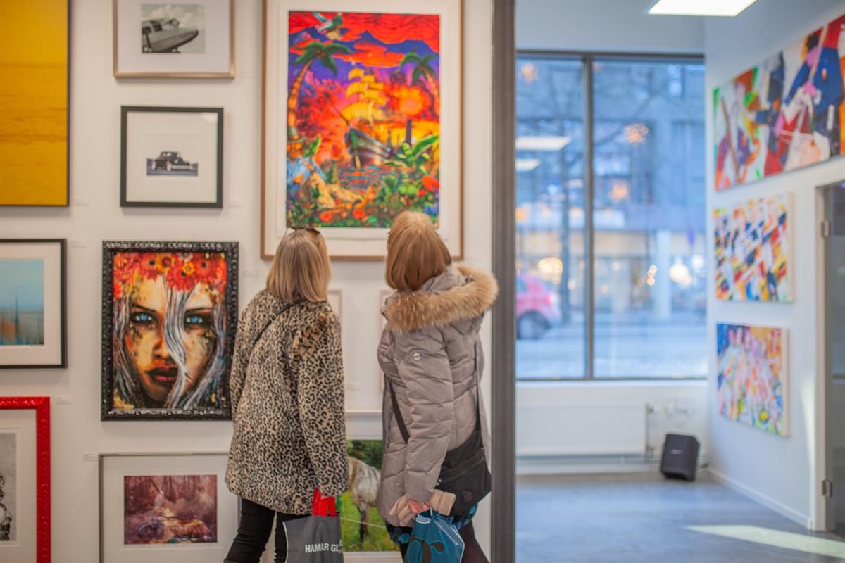 Two people are standing in an art gallery, viewing colorful paintings and prints on the walls, with large windows bringing in natural light.