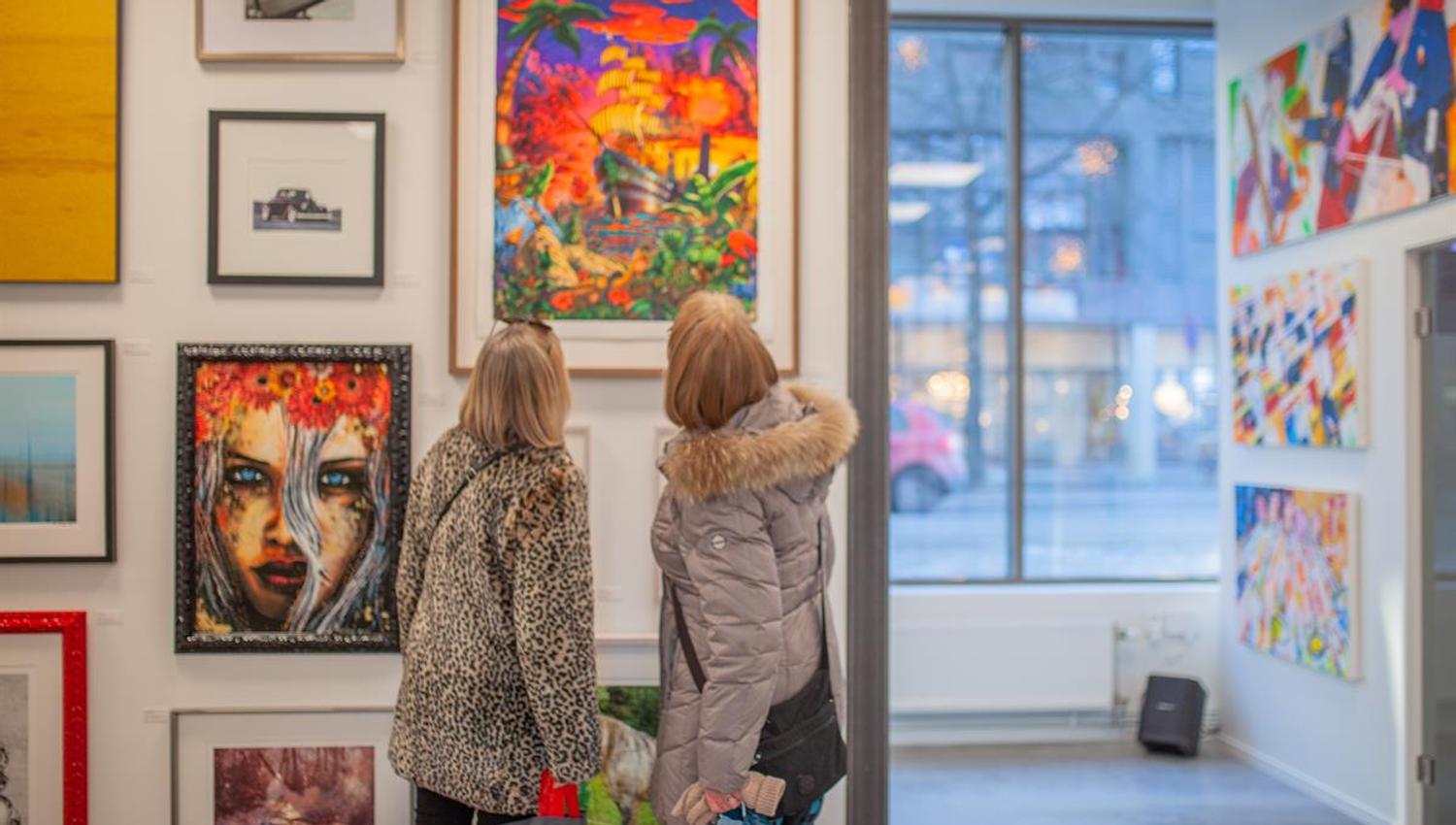 Two people are standing in an art gallery, viewing colorful paintings and prints on the walls, with large windows bringing in natural light.