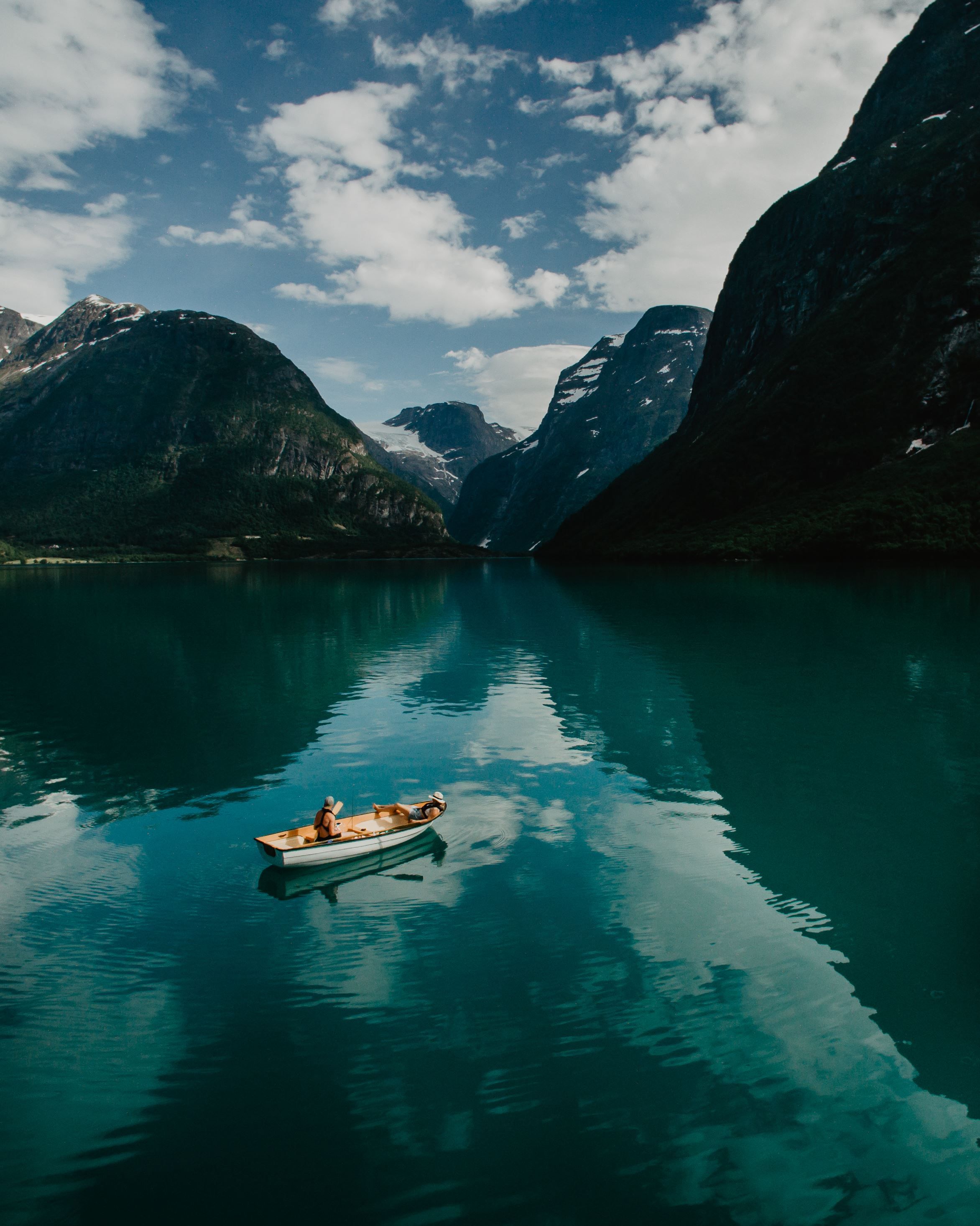 A small rowboat with two people floats on calm turquoise water, surrounded by steep mountains and clouds..