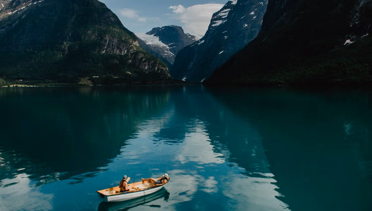 A small rowboat with two people floats on calm turquoise water, surrounded by steep mountains and clouds..