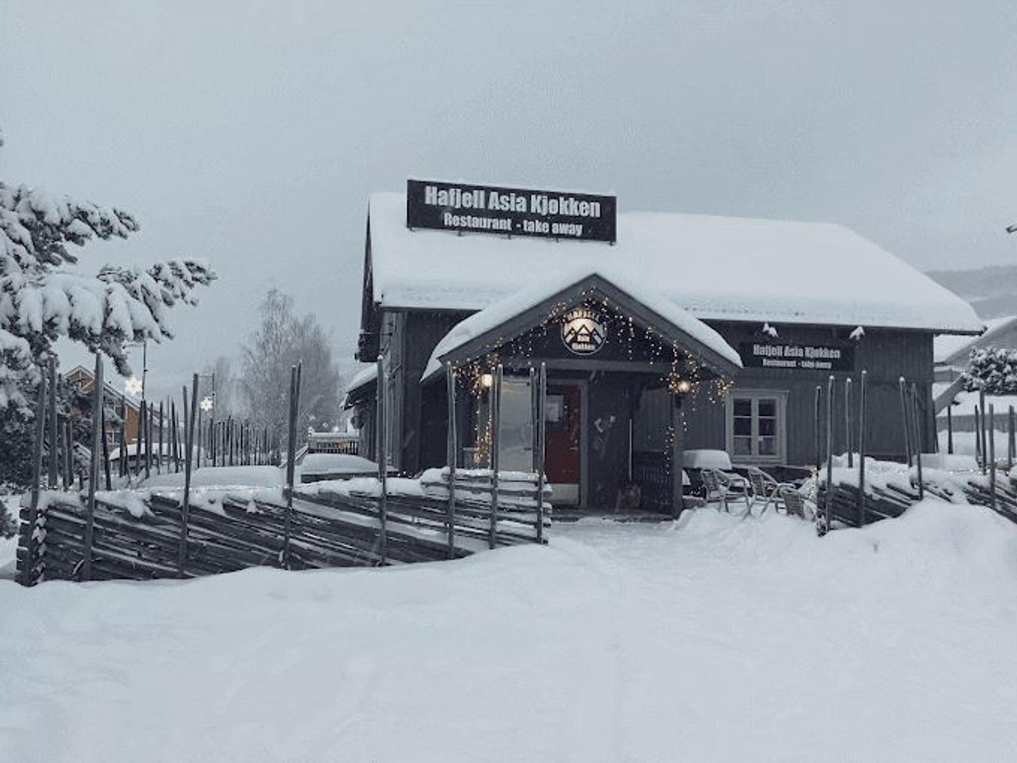 Trehus med skilt og lyslenker, omkranset av gjerder og dyp snø under grå vinterhimmel..