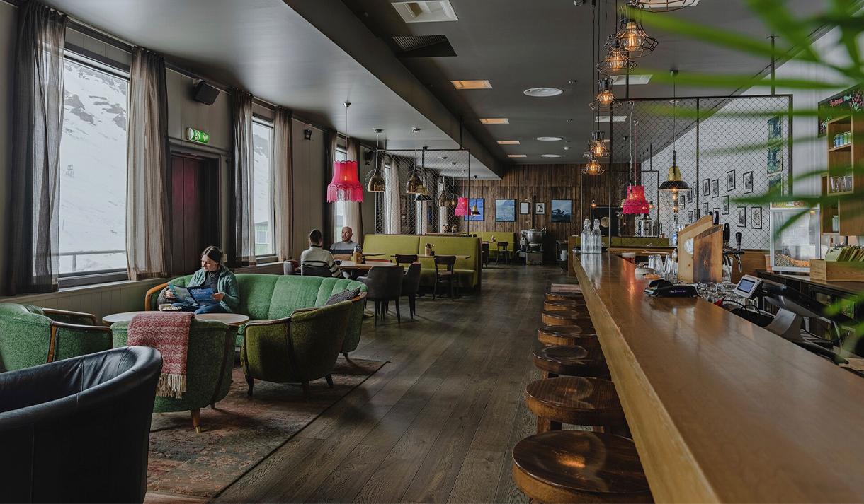En resepsjon og lounge med en gjest som sitter i en sofa i forgrunnen, og restaurantbord med gjester ved et bord i bakgrunnen