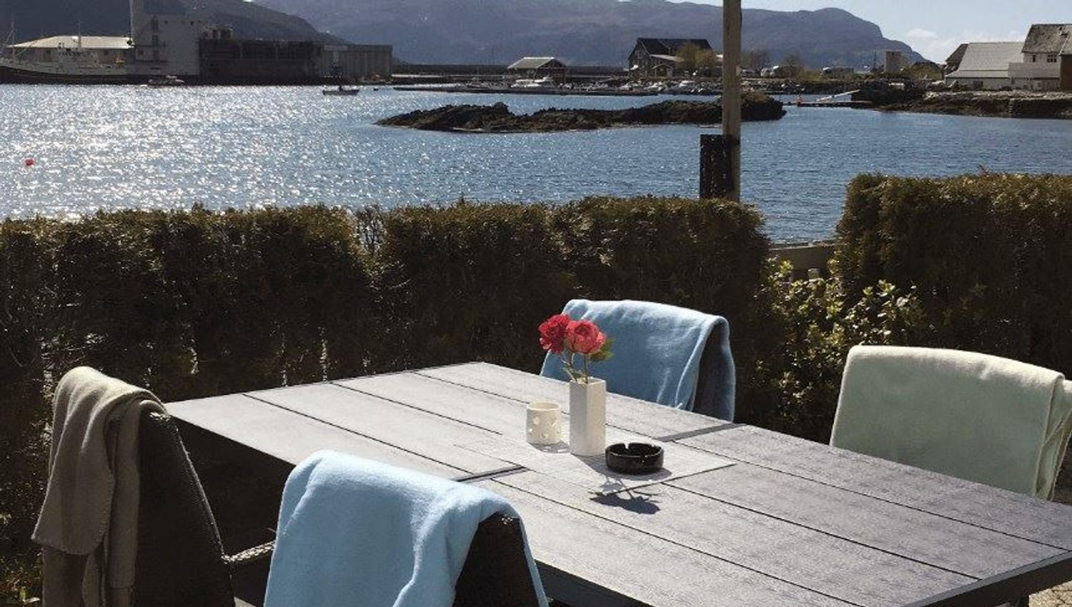Outdoor patio table with chairs and draped blankets, a vase of flowers, and a sunny harbor with mountains beyond..