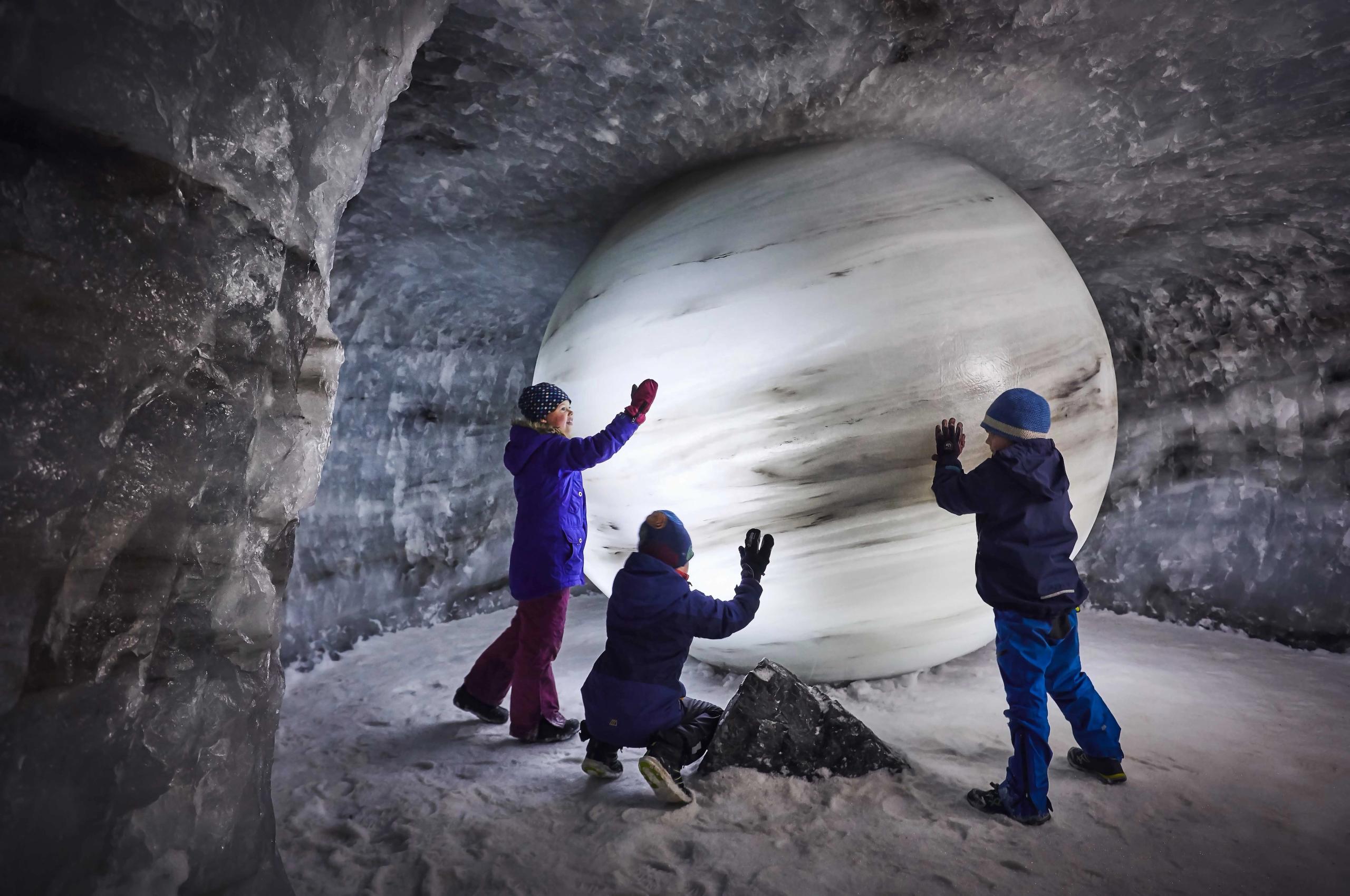 Three children in winter clothing stand and kneel inside an ice cave, facing a large white sphere on snow..