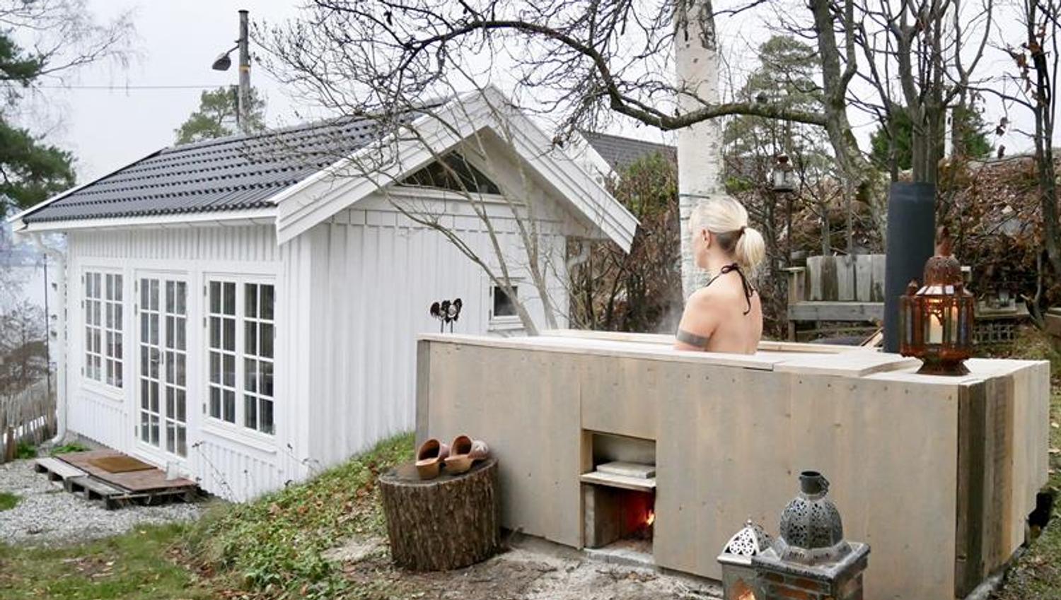 Lite, hvitt tiny house med sovehems og store vinduer, plassert i en grønn hage med trær. person i et badekar foran huset med en terrasse med utsikt mot fjorden.