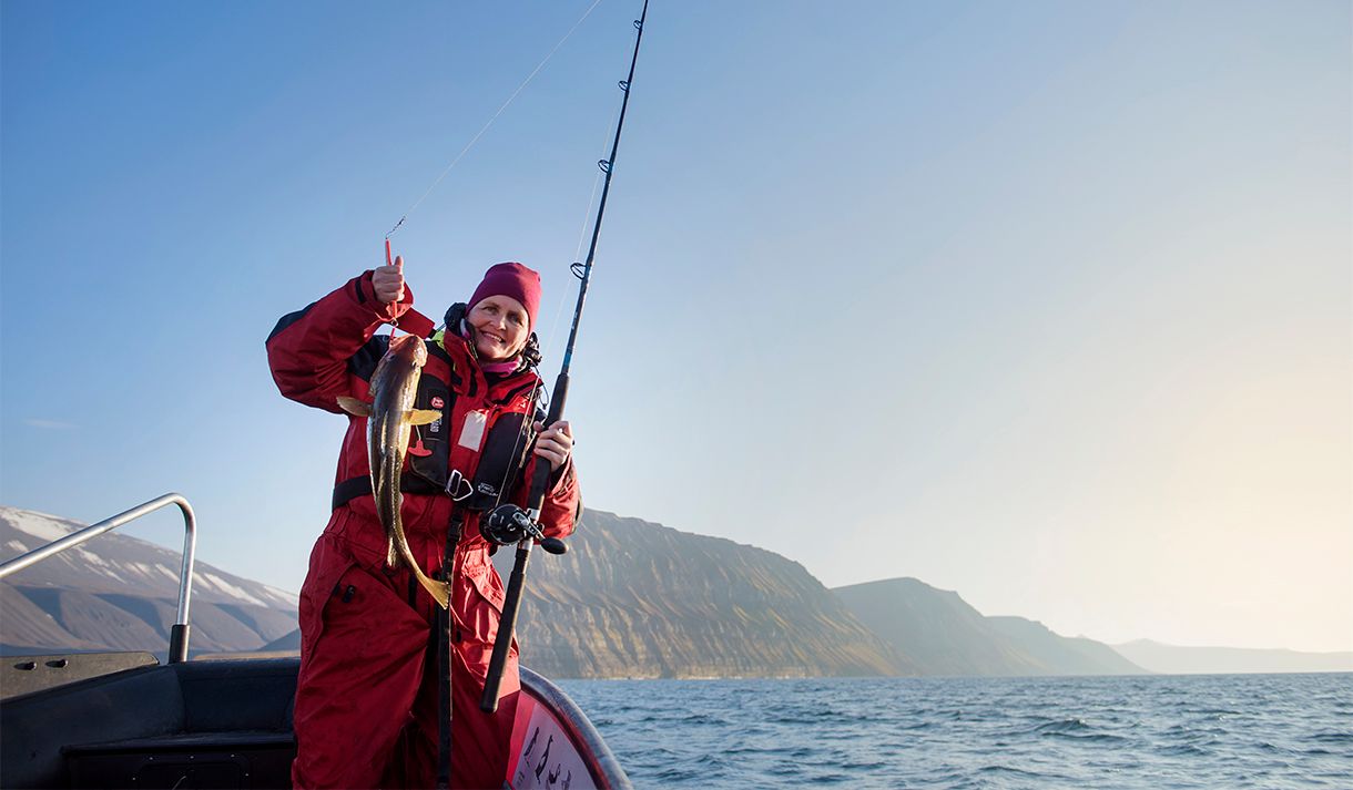 En gjest som står i en RIB båt med en fiskestang og en fanget fisk i forgrunnen, med en åpen fjord og fjell i bakgrunnen
