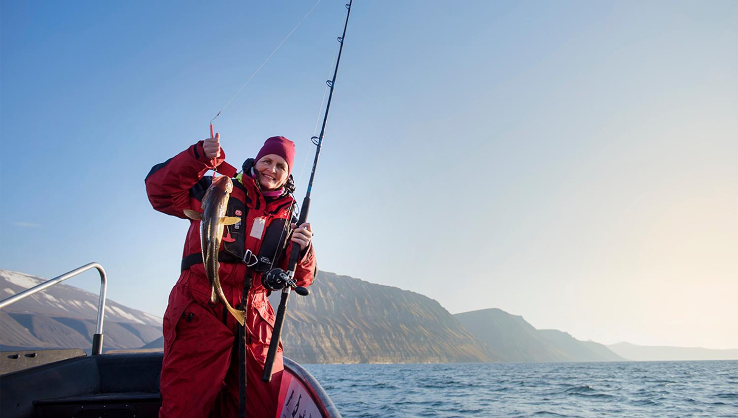 En gjest som står i en RIB båt med en fiskestang og en fanget fisk i forgrunnen, med en åpen fjord og fjell i bakgrunnen