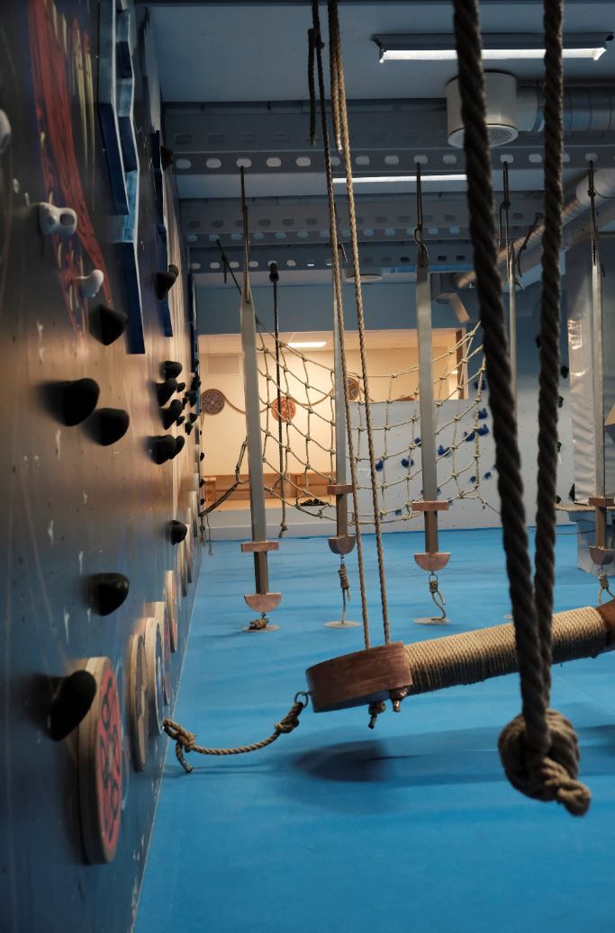 Empty indoor adventure gym with climbing holds, hanging ropes, net obstacles, and a padded blue floor..