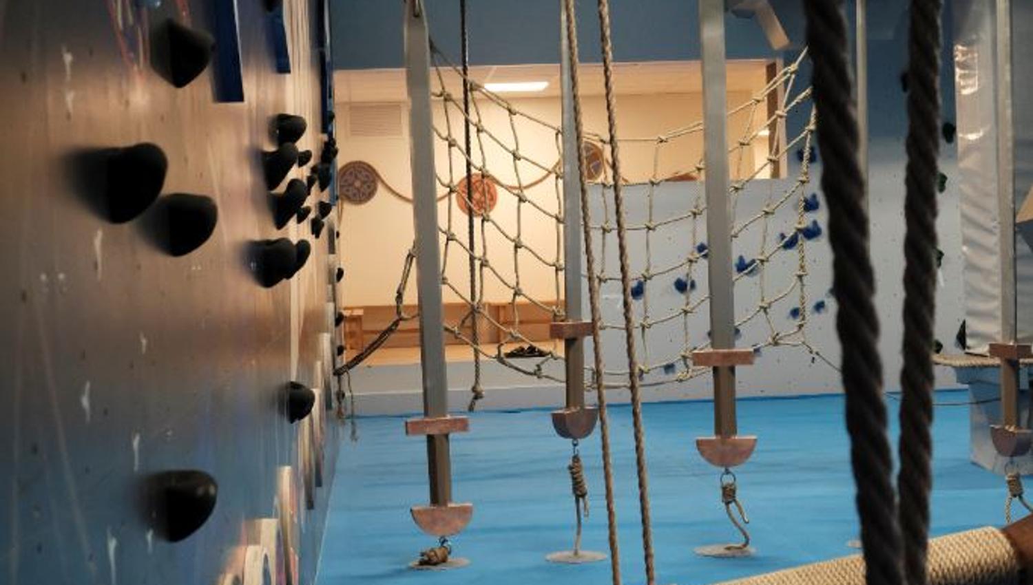 Empty indoor adventure gym with climbing holds, hanging ropes, net obstacles, and a padded blue floor..