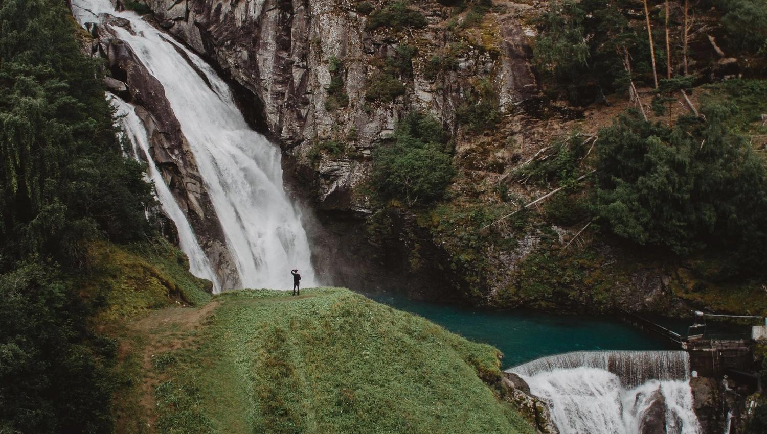 A person stands on a grassy hill beside a tall waterfall dropping into a blue pool, with rocky forested cliffs.