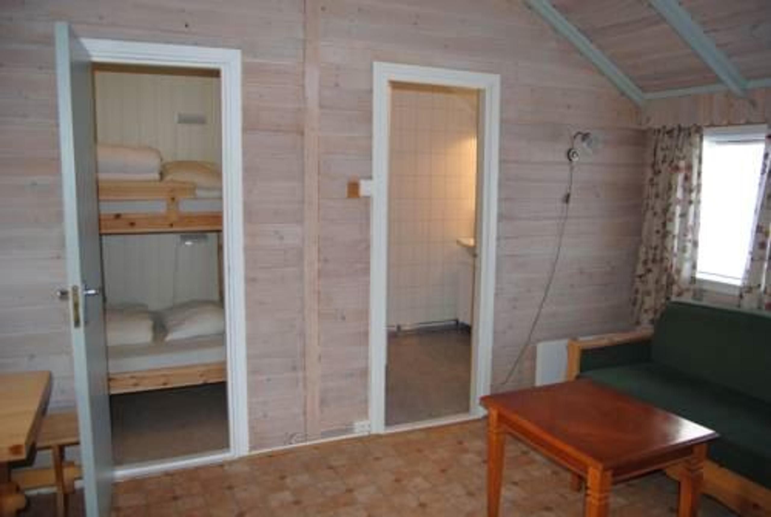 Rustic cabin interior with open door to bunk beds, a bathroom doorway, green sofa, and wooden coffee table.