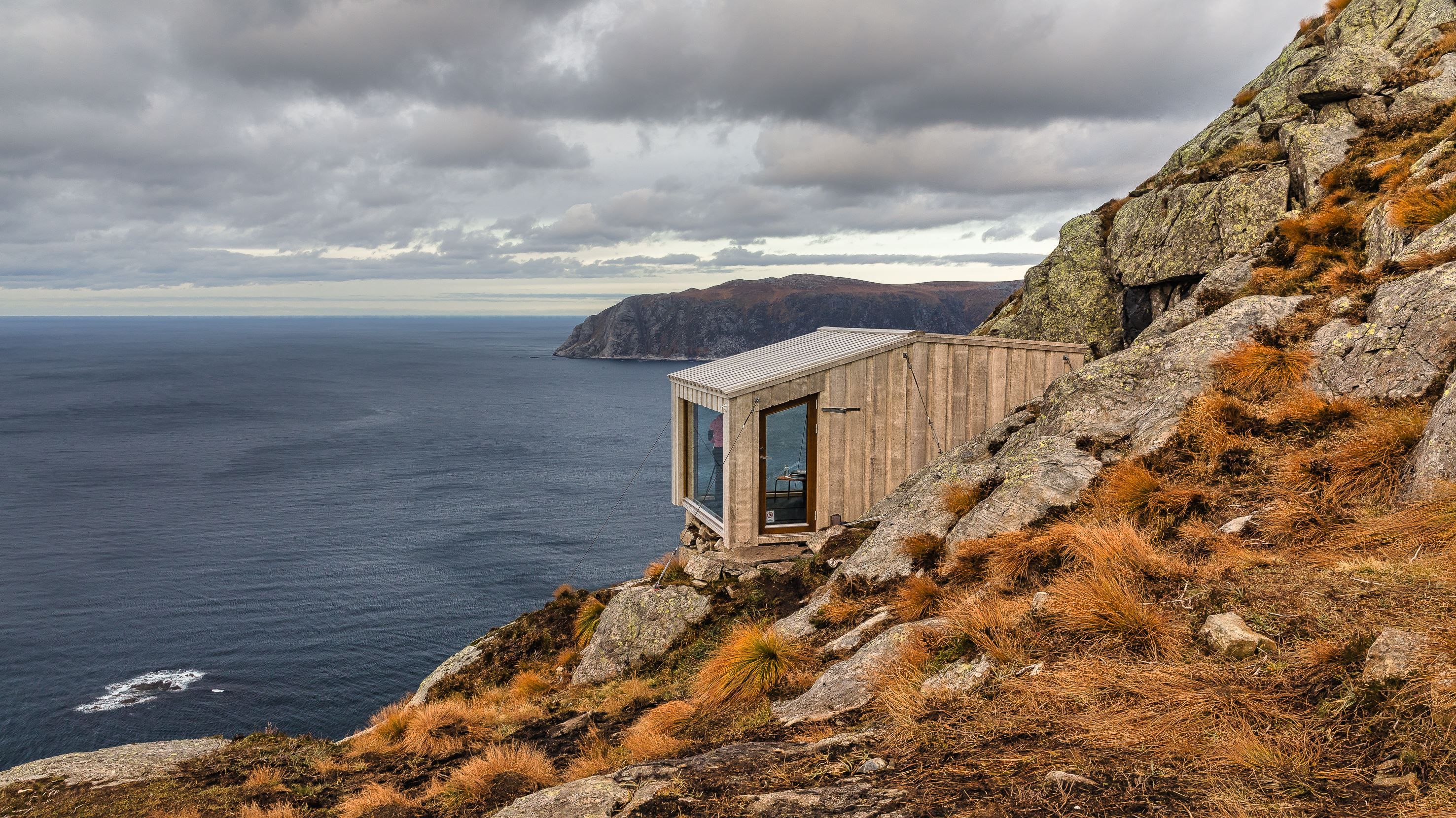 A small wooden cabin clings to a rocky coastal cliff above the ocean, under overcast skies and rugged grass..