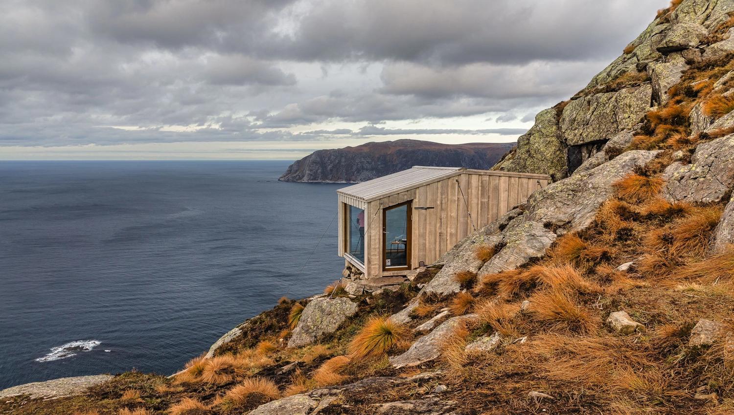 A small wooden cabin clings to a rocky coastal cliff above the ocean, under overcast skies and rugged grass..