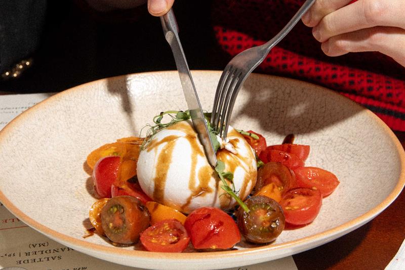 En gjest skjærer i en burrata servert med tomater og dressing på en restaurant.