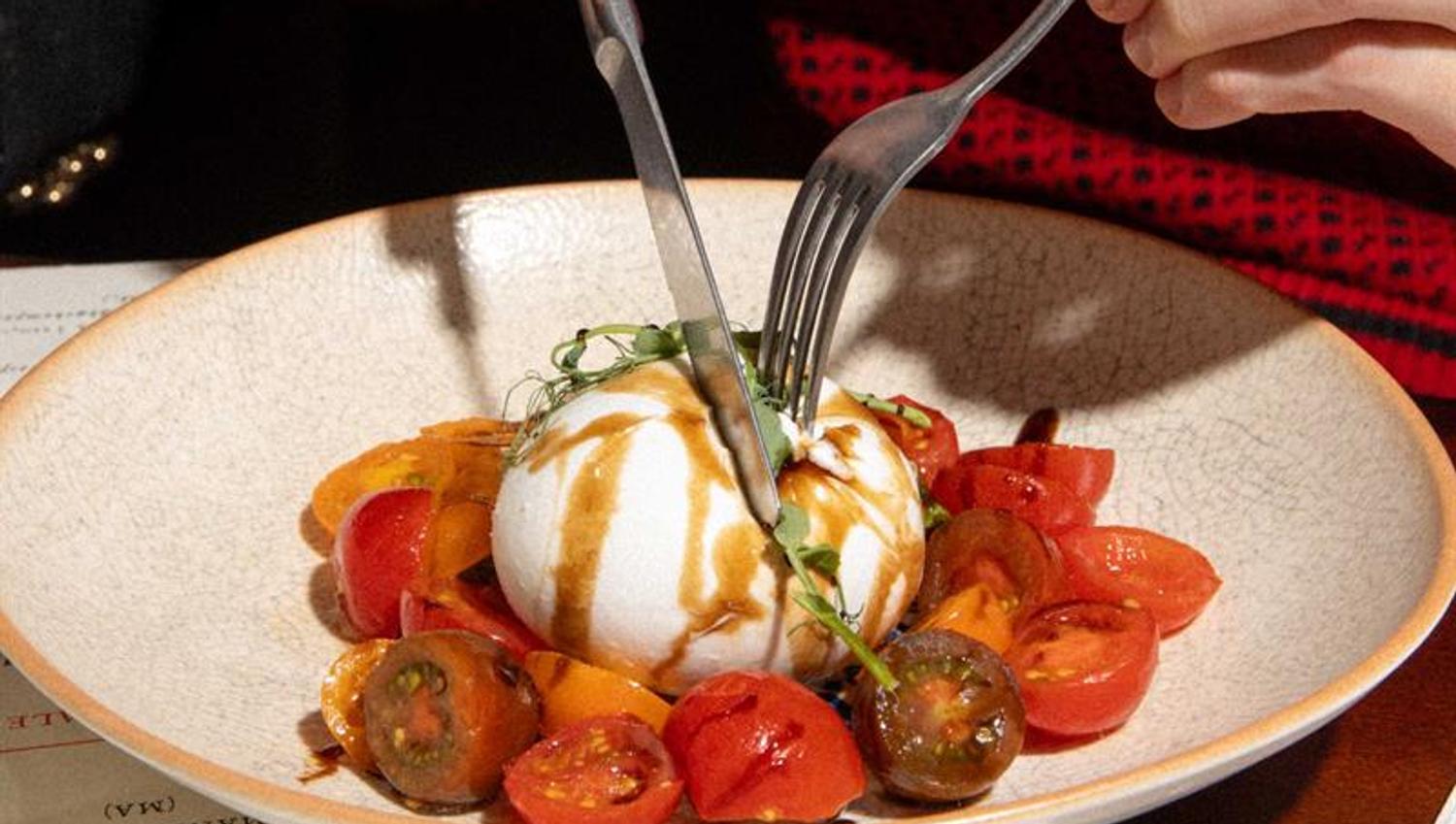 En gjest skjærer i en burrata servert med tomater og dressing på en restaurant.