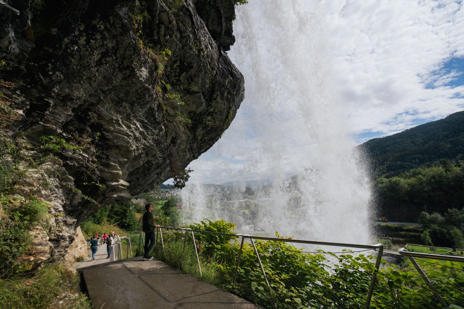 Close-up of visitors walking under Steinsdalsfossen on a dedicated path, with the water cascading powerfully in the background and the Norwegian natur