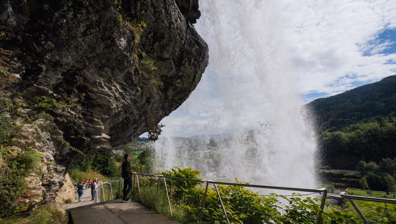 Close-up of visitors walking under Steinsdalsfossen on a dedicated path, with the water cascading powerfully in the background and the Norwegian natur