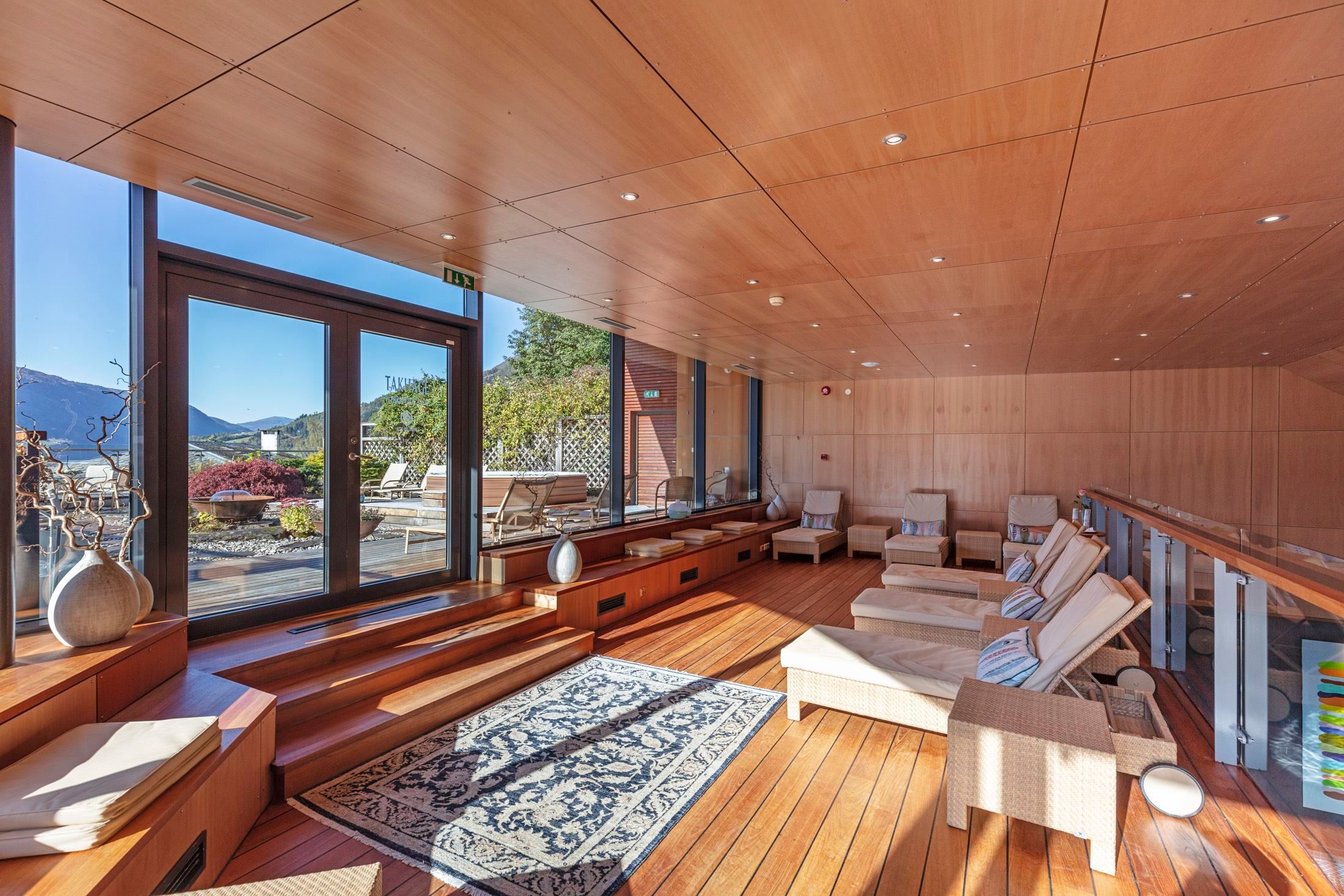 Bright wooden lounge with reclining chairs, rug, and large glass doors opening to a terrace and distant mountains at Alexandra Spa..