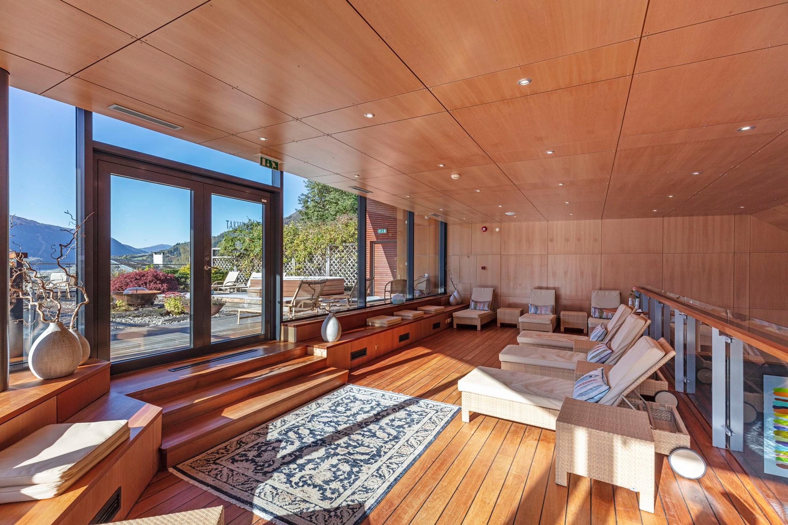 Bright wooden lounge with reclining chairs, rug, and large glass doors opening to a terrace and distant mountains at Alexandra Spa..