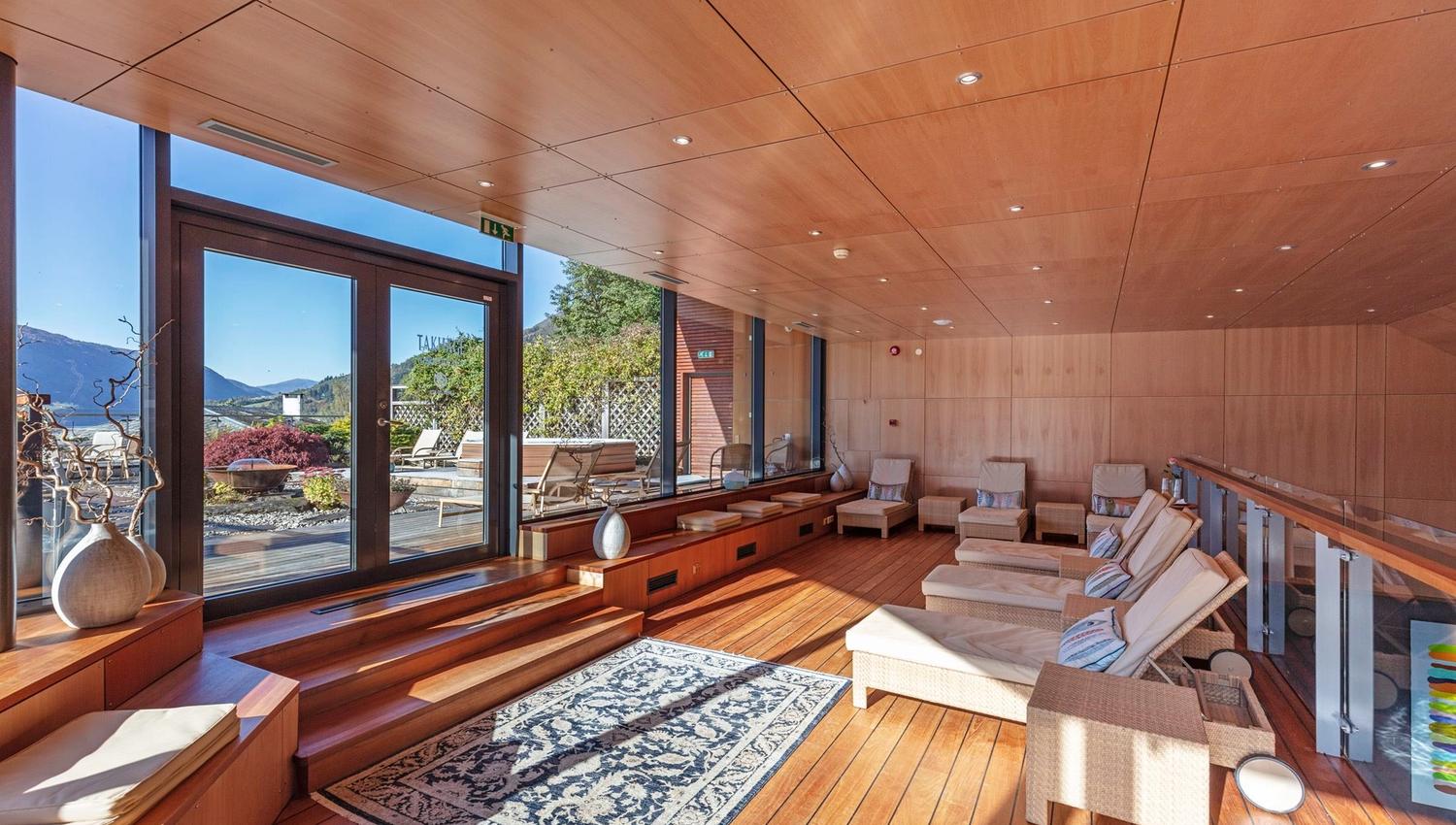 Bright wooden lounge with reclining chairs, rug, and large glass doors opening to a terrace and distant mountains at Alexandra Spa..