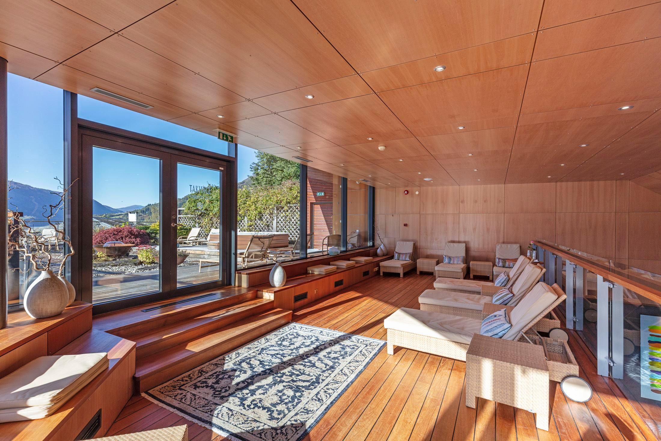 Bright wooden lounge with reclining chairs, rug, and large glass doors opening to a terrace and distant mountains at Alexandra Spa..