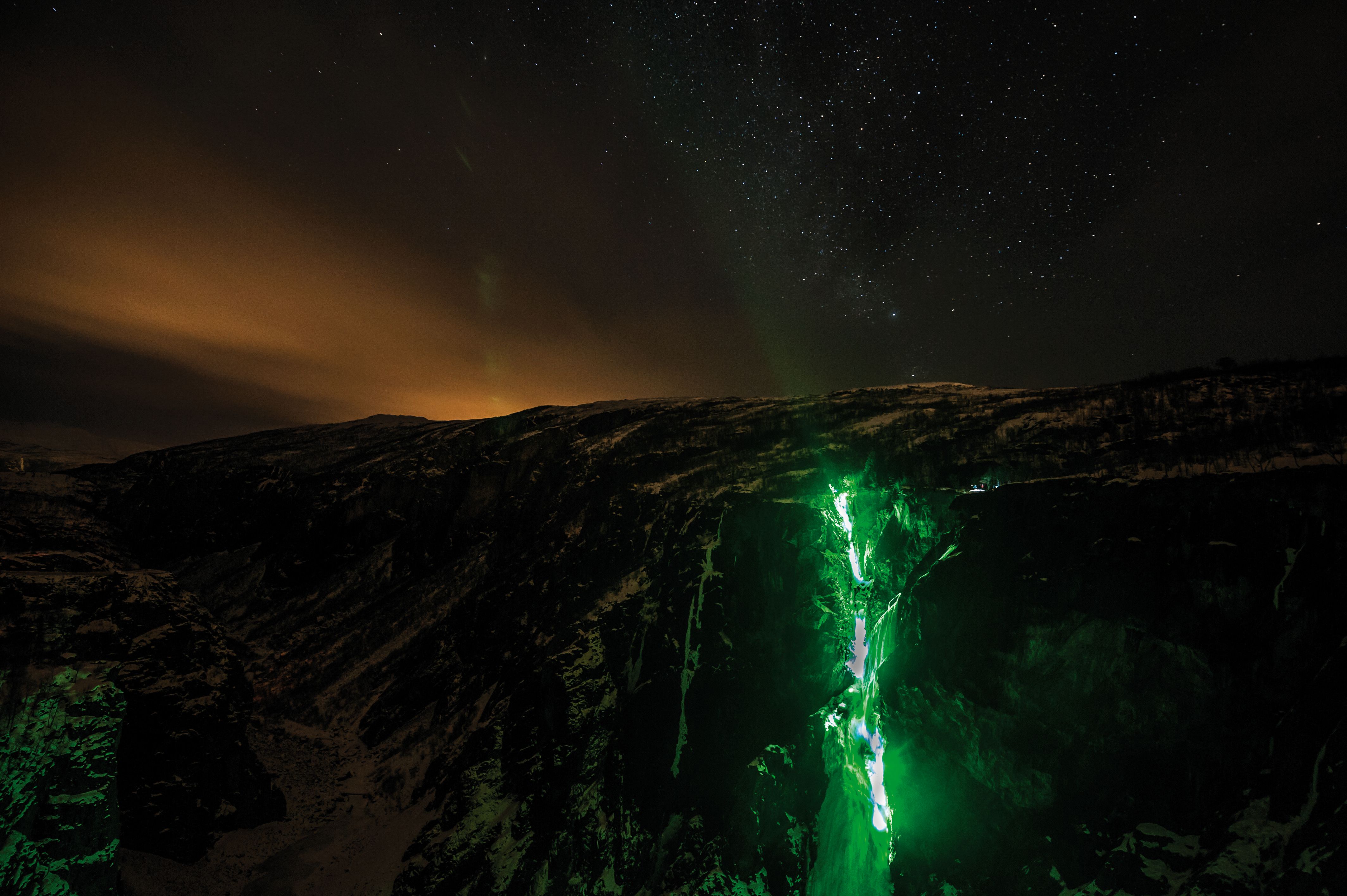 A night time photo of the Tyssvikjo waterfall which is frozen and lit up by green lights.