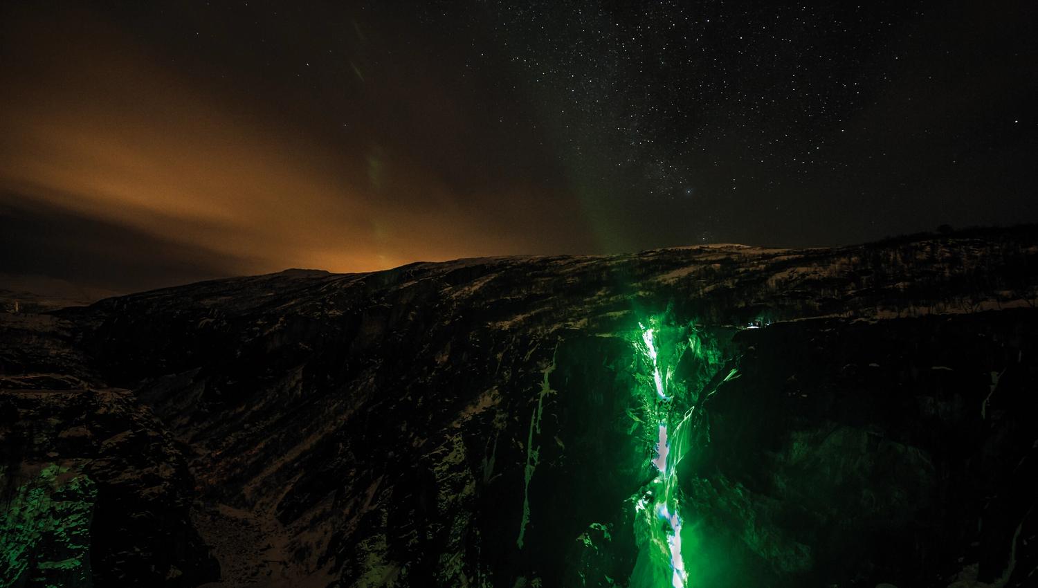 A night time photo of the Tyssvikjo waterfall which is frozen and lit up by green lights.