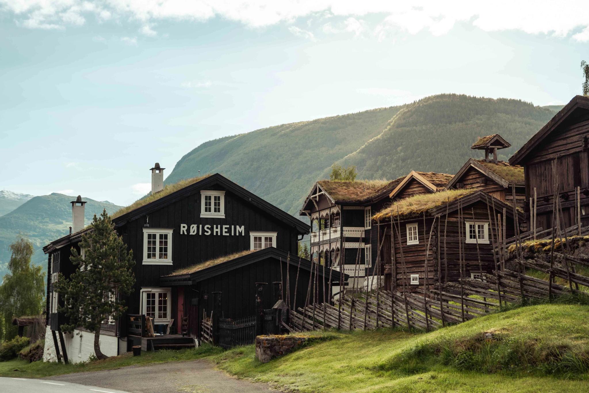 Mørk trebygning med skiltet «RØISHEIM» og fleire lafta hus med torvtak i grøn fjellbygd..