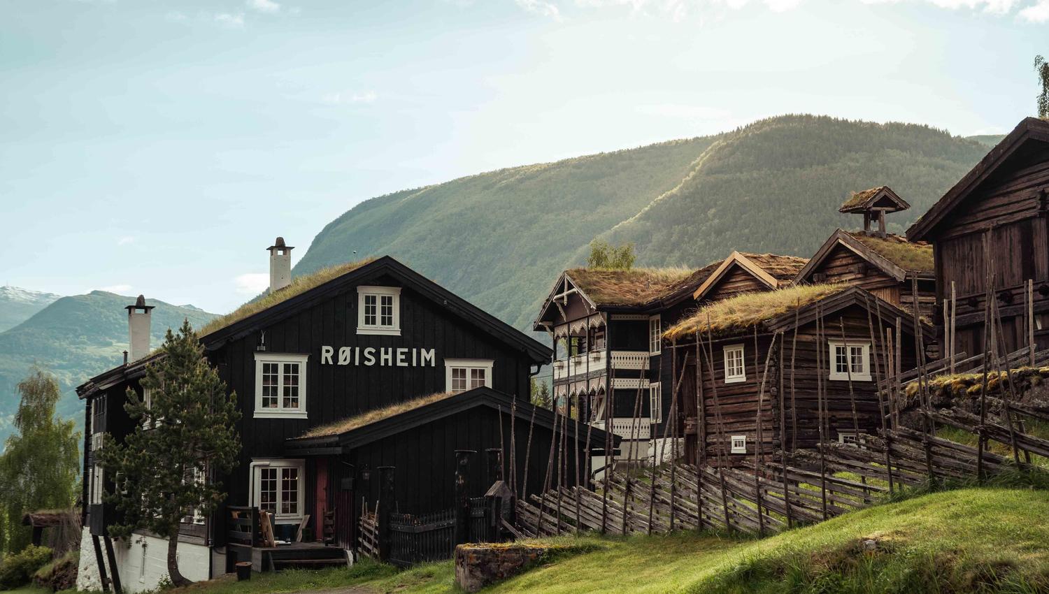 Mørk trebygning med skiltet «RØISHEIM» og fleire lafta hus med torvtak i grøn fjellbygd..